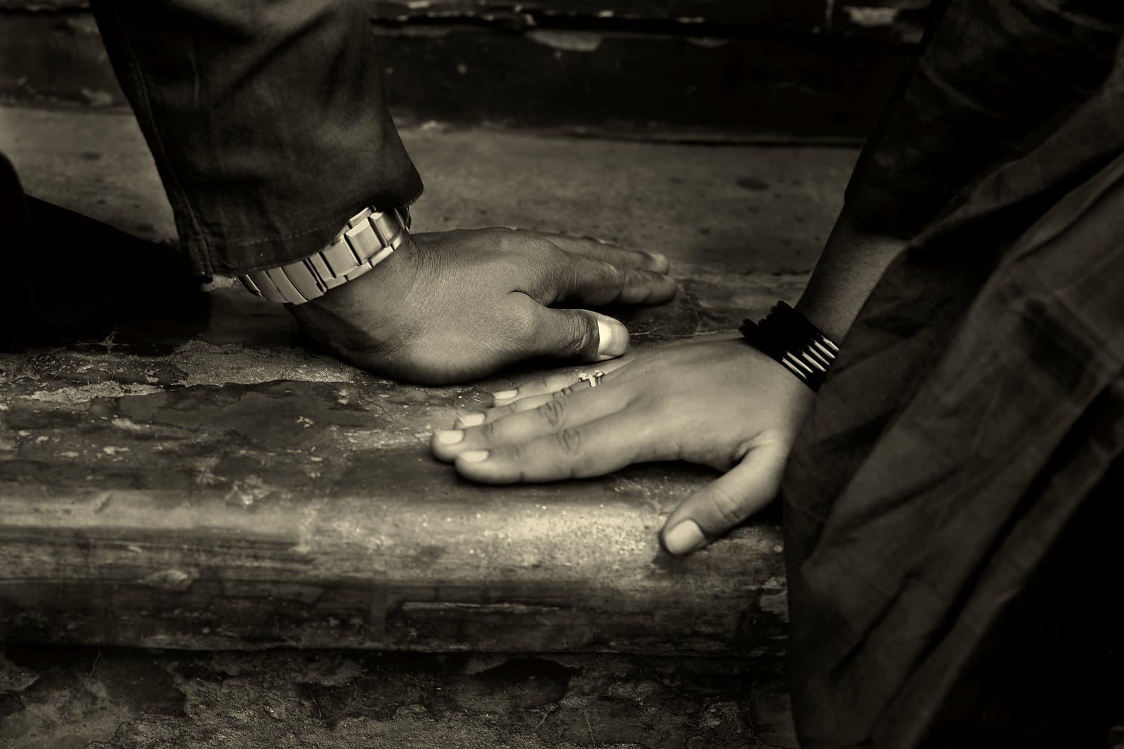 A larger weathered hand resting over a smaller hand on a worn stone step, warm-toned black and white.