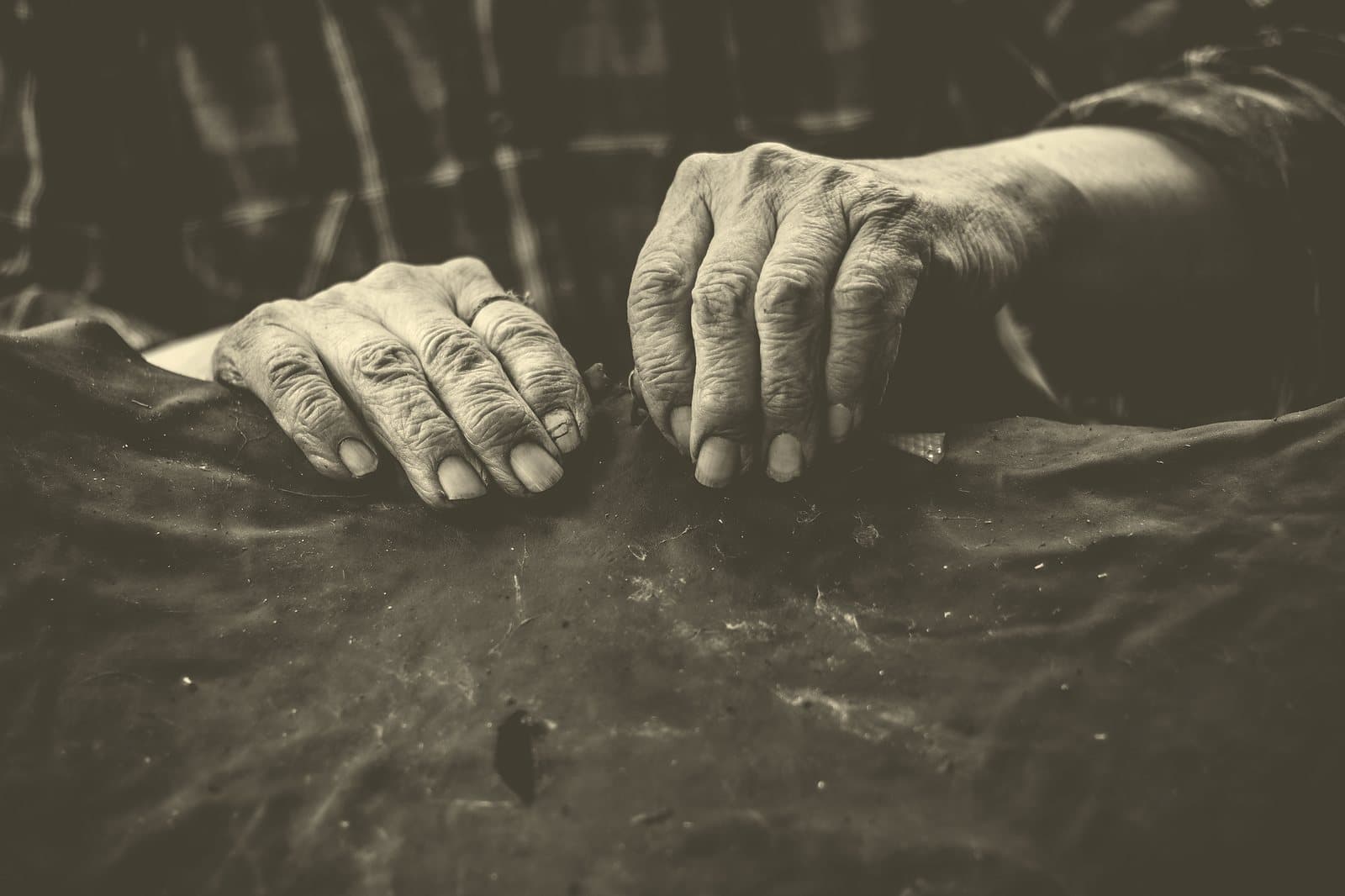 Two deeply weathered elder hands resting on a worn leather work surface, in warm-toned black and white.