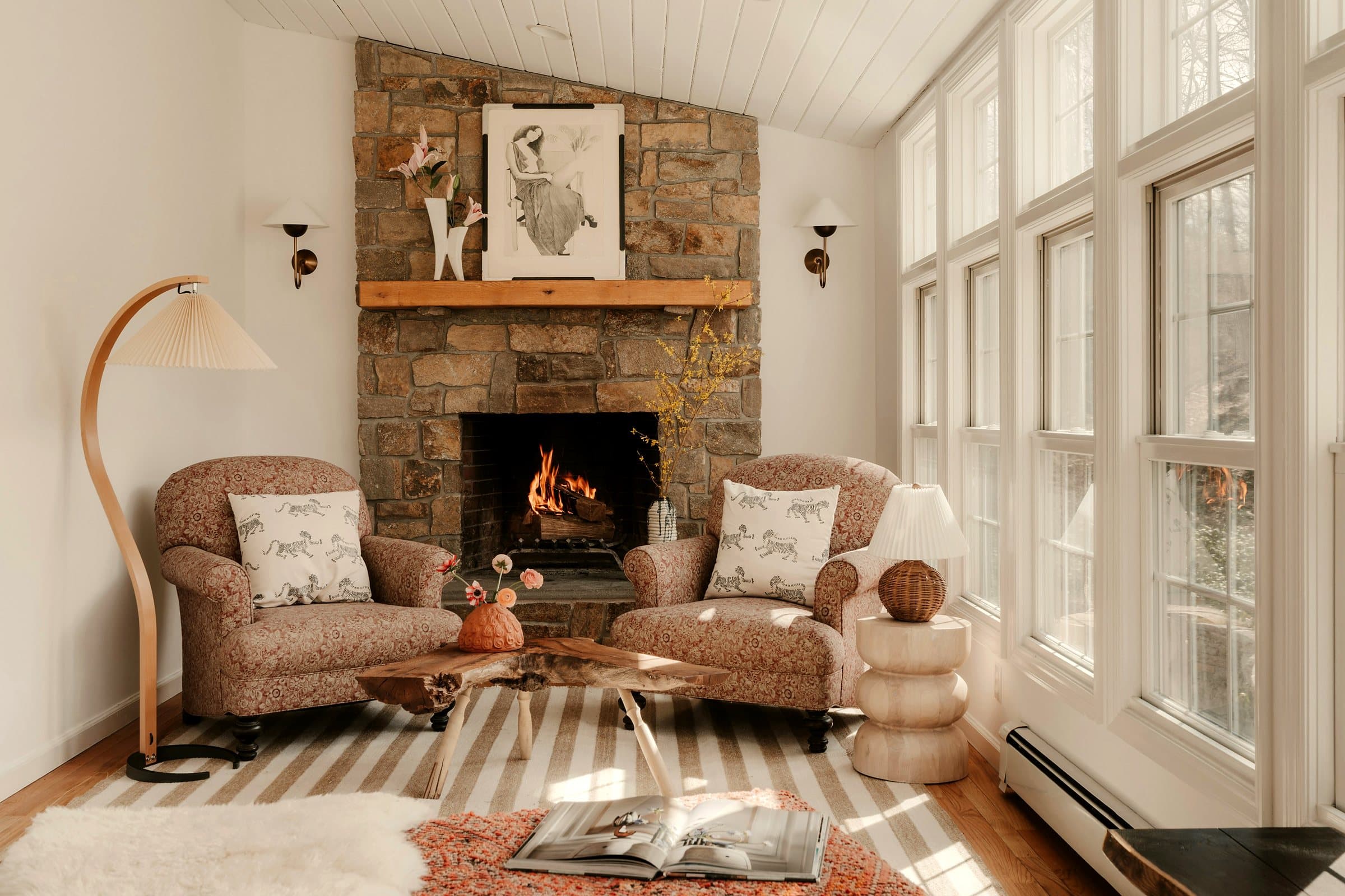 Two upholstered armchairs facing a fieldstone hearth with a lit fire and an oak mantel, in a sun-filled room.