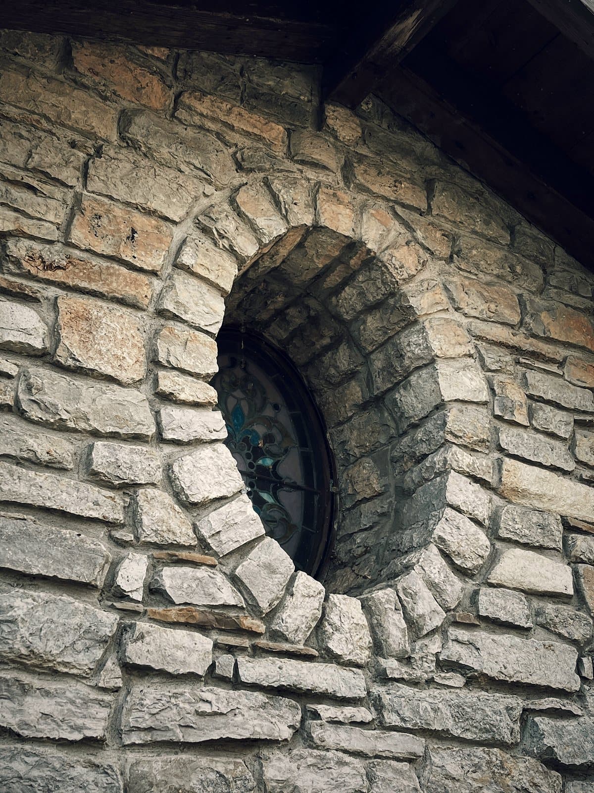 A round stained-glass window set into a stone wall, framed by hand-cut voussoir stones laid in a ring around the opening.